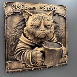 May include: A bronze-colored relief sculpture of a grumpy cat holding a coffee mug. The text "Coffee First, Talk Later" is above the cat. The cat is wearing a robe. The sculpture has a textured, aged appearance.