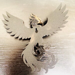 May include: A silver-colored metal phoenix wall hanging with outstretched wings. The phoenix has detailed feathering and a golden head. It is designed to be mounted on a wall and is a decorative item.