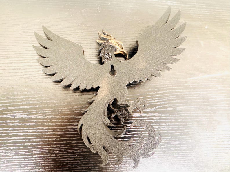 May include: A silver-colored metal phoenix wall hanging with outstretched wings. The phoenix has detailed feathering and a golden head. It is designed to be mounted on a wall and is a decorative item.