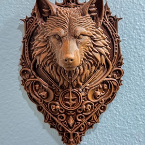 Peut inclure: Décoration murale en forme de tête de loup sculptée en marron. Le loup est centré dans un cadre décoratif en forme de bouclier avec des détails ornés, notamment des crânes et des motifs géométriques. La pièce est fixée sur un mur bleu clair.