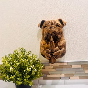 May include: A wooden bulldog wall sculpture, holding a candle. The sculpture is a warm brown color, with detailed carving. The bulldog's expression is serious. The candle is also brown. The sculpture is mounted on a white wall.