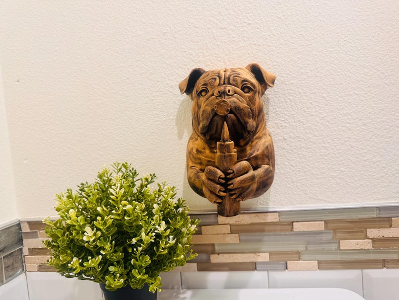 May include: A wooden bulldog wall sculpture, holding a candle. The sculpture is a warm brown color, with detailed carving. The bulldog's expression is serious. The candle is also brown. The sculpture is mounted on a white wall.