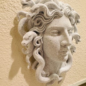K&ouml;nnte beinhalten: Hellgraue Medusa-Kopf-Skulptur mit detaillierten Schlangen und Gesichtsz&uuml;gen. Die Skulptur ist an einer beigefarbenen Wand befestigt und zeigt das komplizierte Design und die Textur des Kunstwerks. Die Schlangen sind um den Kopf gewickelt.
