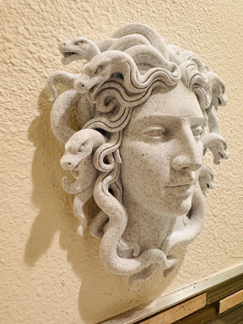 K&ouml;nnte beinhalten: Hellgraue Medusa-Kopf-Skulptur mit detaillierten Schlangen und Gesichtsz&uuml;gen. Die Skulptur ist an einer beigefarbenen Wand befestigt und zeigt das komplizierte Design und die Textur des Kunstwerks. Die Schlangen sind um den Kopf gewickelt.