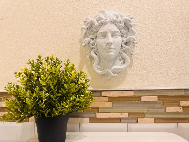 K&ouml;nnte beinhalten: Eine wei&szlig;e Medusa-Kopf-Skulptur ist an einer beigefarbenen Wand angebracht. Die Skulptur hat detaillierte Gesichtsz&uuml;ge und Schlangen als Haare. Eine gr&uuml;ne Topfpflanze steht vor der Wand. Der Topf ist schwarz.
