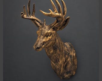 Op de afbeelding: Een wandsculptuur van een hertenkop met een metallic gouden en donkerbruine afwerking. Het hert heeft gedetailleerd gewei, een textuurrijke vacht en een vooruitkijkende blik, vormgegeven als een opgezette kop met een verweerde uitstraling.