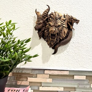 May include: A detailed, brown wall sculpture depicting a mythical Chimera with three heads: a goat, a lion, and a serpent. The sculpture is mounted on a white wall, showcasing intricate details and textures.