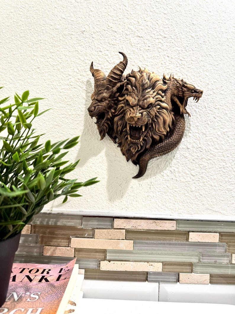 May include: A detailed, brown wall sculpture depicting a mythical Chimera with three heads: a goat, a lion, and a serpent. The sculpture is mounted on a white wall, showcasing intricate details and textures.