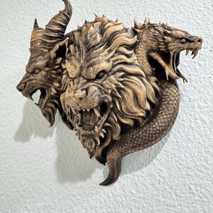 May include: A detailed, bronze-colored wall sculpture depicting a mythical creature with three heads: a goat, a lion, and a serpent. The sculpture has intricate details and is mounted on a textured white wall.
