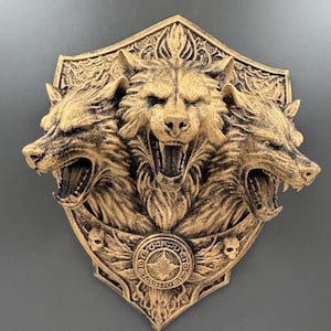 May include: A gold-coloured, three-headed wolf sculpture mounted on a shield-shaped plaque. The wolves are snarling, with detailed fur and open mouths. The plaque features ornate detailing and two small skulls.