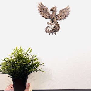 May include: A detailed, brown wooden phoenix wall art piece with outstretched wings is mounted on a white wall. The sculpture has intricate feather detailing and a stylized, regal appearance. A potted green plant sits below.