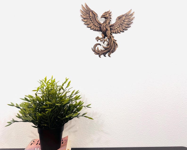 May include: A detailed, brown wooden phoenix wall art piece with outstretched wings is mounted on a white wall. The sculpture has intricate feather detailing and a stylized, regal appearance. A potted green plant sits below.
