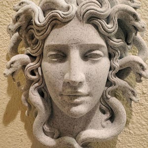 K&ouml;nnte beinhalten: Graue Skulptur von Medusas Gesicht mit geschlossenen Augen und detaillierten Gesichtsz&uuml;gen. Ihr Haar besteht aus zahlreichen Schlangen mit detaillierten Schuppen. Die Skulptur ist an einer strukturierten, hellfarbigen Wand befestigt.
