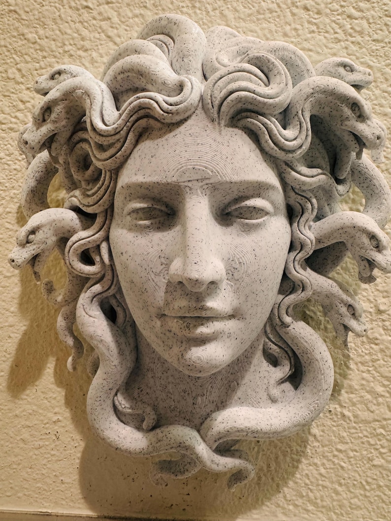 K&ouml;nnte beinhalten: Graue Skulptur von Medusas Gesicht mit geschlossenen Augen und detaillierten Gesichtsz&uuml;gen. Ihr Haar besteht aus zahlreichen Schlangen mit detaillierten Schuppen. Die Skulptur ist an einer strukturierten, hellfarbigen Wand befestigt.