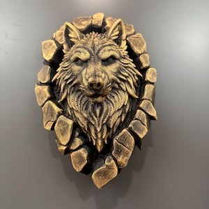 Könnte beinhalten: Detaillierte, goldfarbene Wolfskopfskulptur, die aus einem steinartigen Rahmen hervortritt. Das Fell des Wolfes ist aufwendig detailliert, und die Skulptur hat ein strukturiertes Aussehen. Das Kunstwerk ist auf einer grauen Oberfläche montiert.