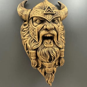 May include: A detailed, golden-colored wall sculpture of a Viking warrior's face. The sculpture features a horned helmet, a long, flowing beard, and an open mouth. The warrior's face is intricately carved with a stern expression.