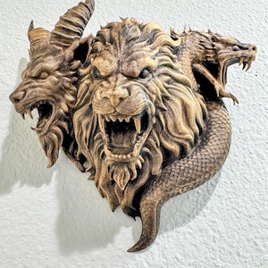 May include: A detailed, bronze-colored wall sculpture depicting a mythical creature with three heads: a lion, a wolf, and a dragon. The sculpture has intricate textures and is mounted on a white wall.