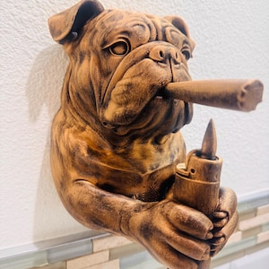 May include: A brown bulldog sculpture with a cigar in its mouth and a lighter in its hand. The sculpture is mounted on a white wall with a decorative tile border. The bulldog has detailed fur and facial features.