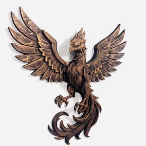 May include: A bronze-colored phoenix wall sculpture with outstretched wings. The bird has detailed feathering and a fierce expression. The sculpture is mounted on a white wall, showcasing the intricate design and texture of the artwork.
