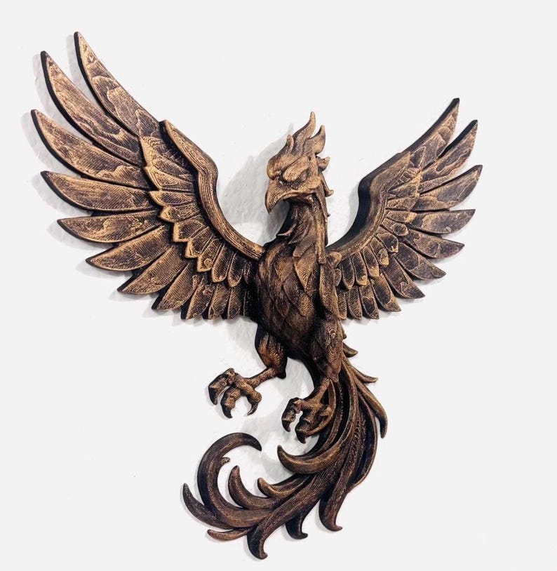 May include: A bronze-colored phoenix wall sculpture with outstretched wings. The bird has detailed feathering and a fierce expression. The sculpture is mounted on a white wall, showcasing the intricate design and texture of the artwork.
