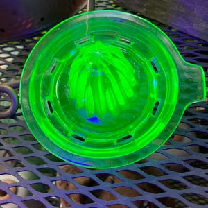 May include: A vibrant, translucent neon green glass citrus juicer featuring a central reamer with vertical ridges, a circular basin with drainage slots, and a small handle tab on the side. The glass glows with a bright, intense fluorescent hue.