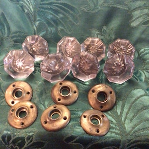 May include: A collection of clear, faceted glass door knobs and aged brass backplates. The knobs have an octagonal shape with a starburst design. The backplates are round with multiple screw holes. The items are arranged on a green patterned fabric.