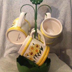 May include: A green metal mug holder with a flower design on top. Four white mugs with yellow and green floral designs are hanging from the holder. The mugs are sitting on a green base.