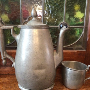 May include: A vintage pewter teapot with a matching cup. The teapot has a rounded body, a curved spout, and a lid with a decorative finial. The cup has a small handle. Both pieces are displayed on a wooden surface in front of a stained glass window.