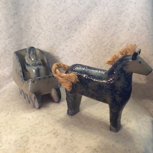 May include: A ceramic horse pulling a sled. The horse is dark blue with a textured finish and a tan mane and tail. The sled is light blue and white, with a figure inside. The horse is connected to the sled with a rope.