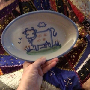 May include: Oval ceramic serving dish with a blue rim and a whimsical cow illustration. The cow is depicted in blue with brown spots, standing on a green patch of grass under a blue cloud. The dish is held by a hand.
