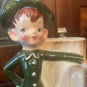 May include: A ceramic figurine of a green-clad elf with a green hat, holding a clear glass orb. The elf has a rosy complexion and brown hair. The figurine is standing next to a tree trunk.
