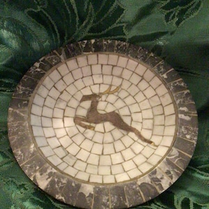 May include: A round, decorative dish made of mosaic tiles. The dish features a stylized deer design in brown and gold against a white background. The outer rim of the dish is made of dark gray stone.