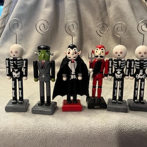 May include: A collection of Halloween-themed nutcrackers. The figures include a skeleton, Frankenstein's monster, a vampire, and a devil. Each figure has a silver wire clip attached to the top. The bases are gray or red and marked with "2011".