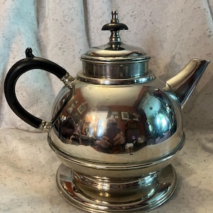 May include: A silver-colored teapot with a black handle and lid finial. The teapot has a round body and sits on a matching base. The polished surface reflects the surrounding environment.
