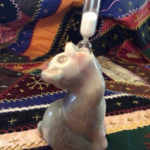 May include: A gray ceramic cat-shaped hourglass with a clear glass top filled with white sand. The cat is seated, with its head turned to the side. The hourglass is set against a colorful patchwork quilt.