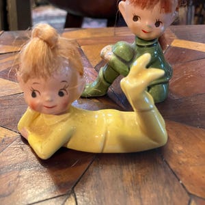 May include: Two ceramic figurines of children with painted faces and hair made of yarn. The figurine on the left is lying down and wearing a yellow outfit. The figurine on the right is sitting with its legs crossed and wearing a green outfit.