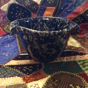 May include: A dark blue and cream speckled ceramic bowl with a wide, fluted rim. The bowl is set against a patchwork quilt with various colors and embroidered designs. The bowl's shape suggests it could be used for serving or display.
