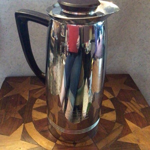 May include: A tall, shiny chrome coffee server with a black handle and a dark brown lid. The server is cylindrical with a spout and sits on a wooden table with a starburst design. The server reflects the surrounding environment.