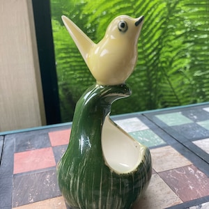 May include: A ceramic bird figurine with a yellow body and green and white striped base. The bird is perched on the base, which has a small opening in the center.