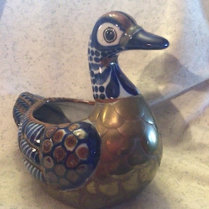 May include: A ceramic duck planter with blue and white painted details and a gold metallic finish. The duck has a round body and a long neck with a small beak.