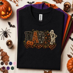 Könnte beinhalten: Ein schwarzes Kurzarm-T-Shirt mit dem Wort DADDY in großen, dekorativen Buchstaben, die mit Halloween-Mustern wie Kürbissen, Geistern und Spinnenweben gefüllt sind. Das Design enthält einen markanten orangefarbenen Kürbis und ein Karomuster.