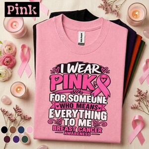 May include: A light pink short-sleeved t-shirt featuring the black and pink text I WEAR PINK FOR SOMEONE WHO MEANS EVERYTHING TO ME BREAST CANCER AWARENESS and a pink ribbon graphic. The shirt is displayed with other folded shirts in black and dark colors.