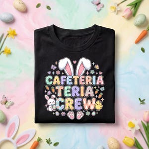 May include: Black t-shirt with the words "CAFETERIA CREW" in pastel colors, decorated with bunny ears, eggs, carrots, butterflies, and a chick. The shirt is folded and ready to wear.