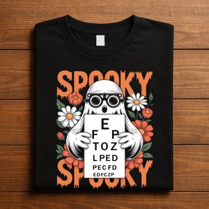 Puede incluir: Camiseta negra con un diseño temático de Halloween. El gráfico presenta un fantasma blanco con gafas que sostiene una tabla optométrica. La palabra "SPOOKY" está escrita en naranja arriba y abajo del fantasma, con flores naranjas y blancas.