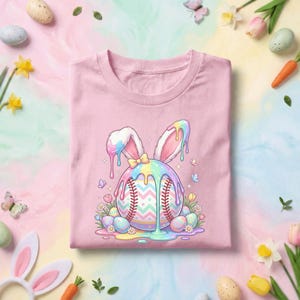 May include: A light pink t-shirt featuring a baseball with bunny ears and colorful pastel dripping paint. The baseball has a zig-zag pattern and red stitching. Easter eggs and flowers surround the baseball. A bow is on the bunny ears.