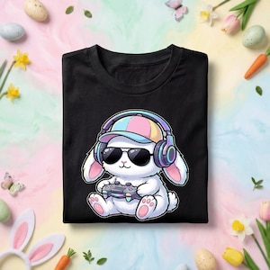 Puede incluir: Camiseta negra con un conejo de dibujos animados con gafas de sol, un sombrero colorido y auriculares. El conejo sostiene un mando de videojuegos. El diseño tiene una combinación de colores pastel, perfecto para Pascua o jugadores.