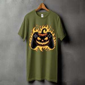 Op de afbeelding: Olijfgroen t-shirt met een Halloween-thema. Het ontwerp toont een zwarte gamecontroller met een pompoengezicht in het midden, omringd door oranje en gele vlammen. Het t-shirt hangt aan een houten hanger.