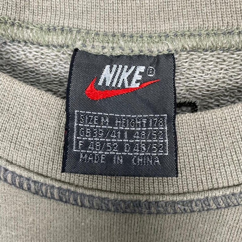 May include: Close-up of a gray sweatshirt with a Nike tag. The tag features the Nike logo in red and white, along with size and manufacturing information. The text "MADE IN CHINA" is visible.