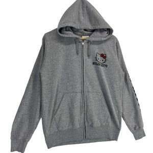 May include: A gray zip-up hoodie featuring a Hello Kitty graphic on the chest. The graphic includes Hello Kitty's face with a red bow and the words "Hello Kitty" below. The hoodie has a drawstring hood and a front pocket.