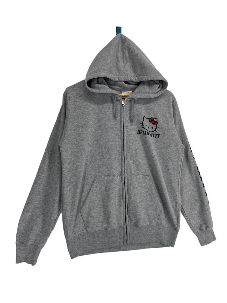 May include: A gray zip-up hoodie featuring a Hello Kitty graphic on the chest. The graphic includes Hello Kitty's face with a red bow and the words "Hello Kitty" below. The hoodie has a drawstring hood and a front pocket.